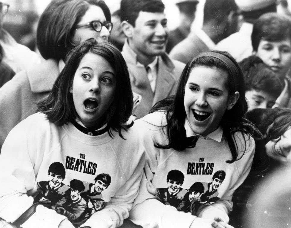 Two excited girls in Beatles sweatshirts, amongst a crowd of fans in New York, welcoming the group as they arrive at the airport.