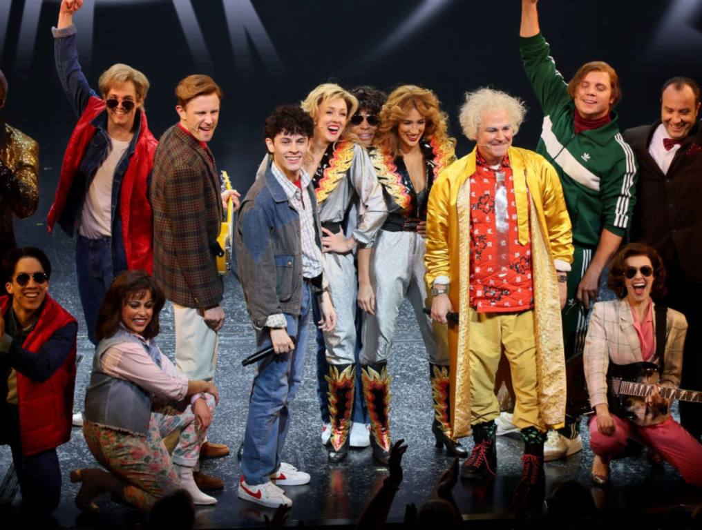 The cast of Back To The Future The Musical stands on stage smiling at the crowd. They are all dressed in the 80's attire from the film.