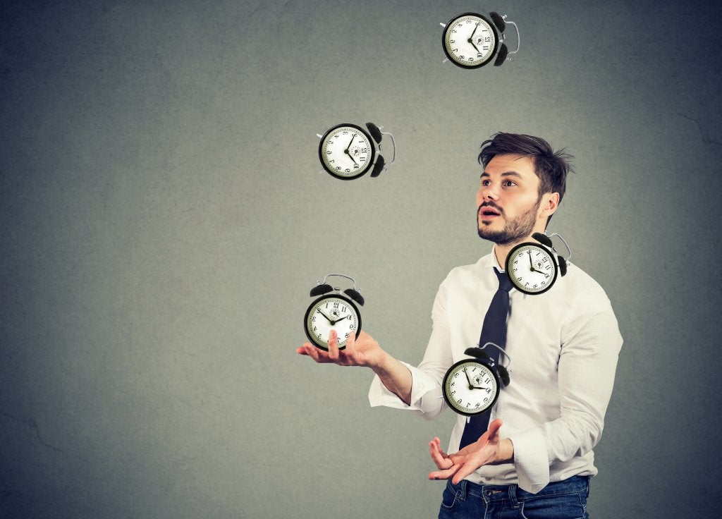 Young brown-hair man with facial hair is wearing a white shirt and patterned tie and juggling multiple analog alarm clocks.