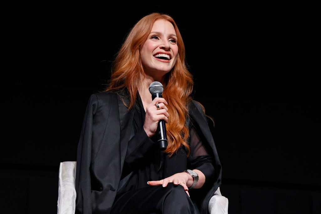 Actress Jessica Chastain sits in a white chair wearing all black and holding a microphone as she smiles at an audience offscreen.