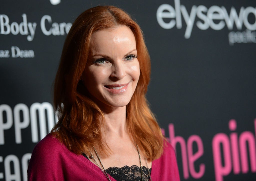 Actress Marcia Cross smiles in a burgundy top as she poses in front of a banner with her red hair down at shoulder length.