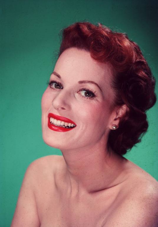 Actress Maureen O'Hara smiles in a headshot against a green backdrop. She has her red hair back and is wearing red lipstick.