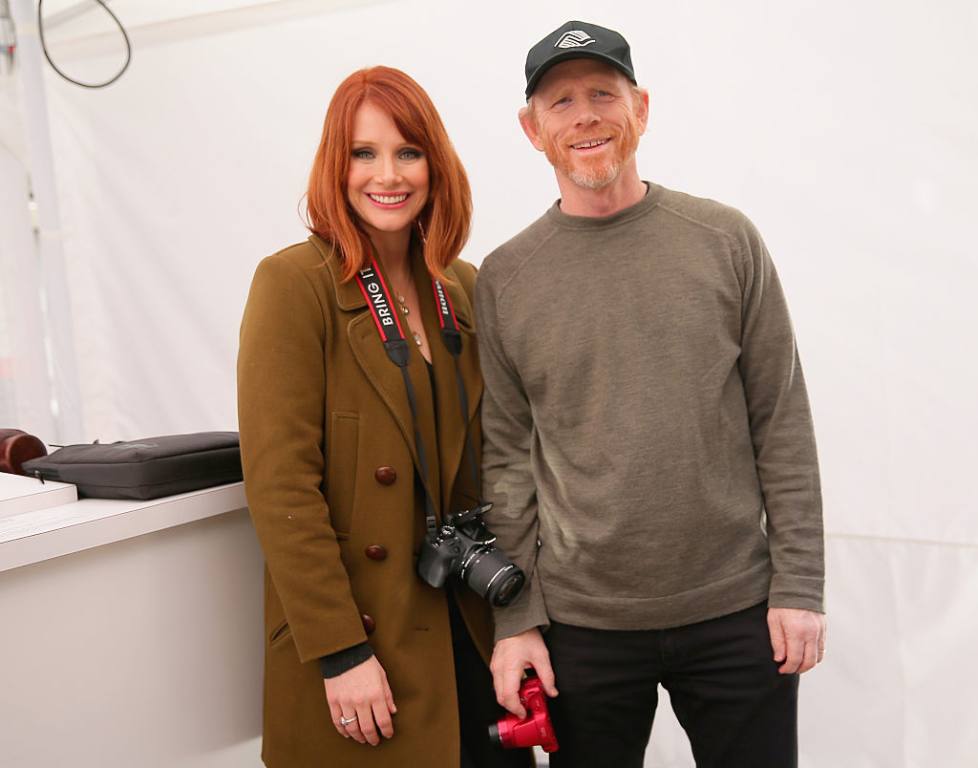 Actress Bryce Dallas Howard poses next to her father, director Ron Howard in front of a white wall. They are both carrying cameras and smiling.