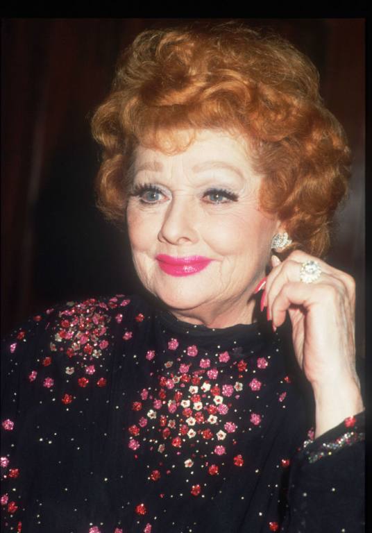 Actress Lucille Ball smiles in a black top with purple dots. Her hair is red and short.