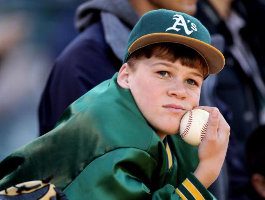 The Oakland A's are proposing a very expensive move to Las Vegas. Photo of a young Oakland A's fan or ball boy, watching the game with a sad look on his face. He is wearing a green Oakland A's baseball cap with a yellow bill. He is also wearing the green warm-up jacket. He is holding a baseball in his right hand up to his chin while he watches the gaem.