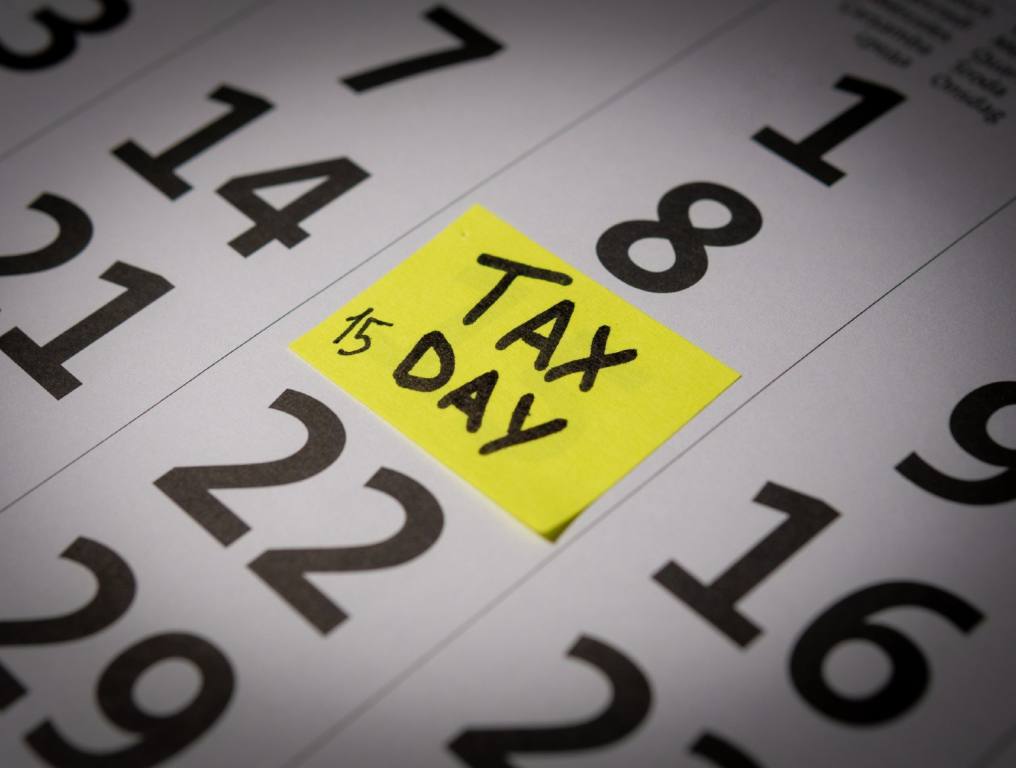 Income Tax Day falls on April 15th most years. Close-up photo of a calendar with black numerals on a white background. There is a yellow post-it note stuck over the number 15 on the calendar. Someone has handwritten the words "Tax Day" in black magic marker on the post-it note. They've also written the number 15 in the bottom left-hand corner of the note.