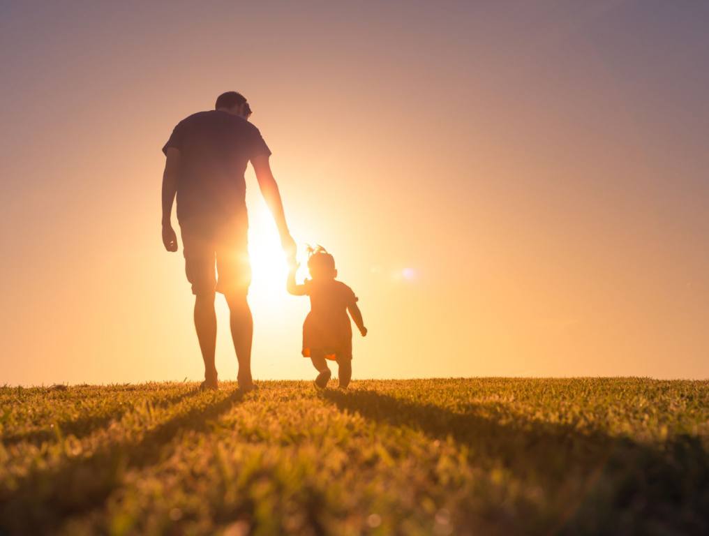 A photo representing Father's Day, with a Dad and his toddler son or daughter walking through a grass field towards a golden sunset.