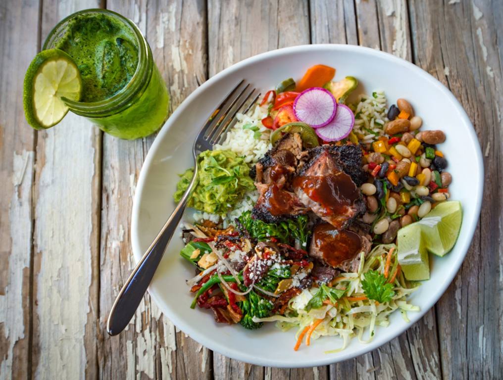 Photo of he BBQ Mexicana "Brisket Lover Bowl." Photo shows chunks of smoked brisket in the middle of an oblong, white, low-rimmed bowl. Surrounding the brisket, going counter-clockwise around the bowl starting at the bottom is Mexican slaw, two lime wedges, a bean salad, white rice topped with colorful chopped peppers and sliced Mexican radishes, a spoonful of guacamole, and a vegetable medley topped with white cheese crumbles. There is a silver fork placed inside the bowl on the left side of the dish. To the left of the bowl is a refreshing green drink in a mason jar. A lime wedge has been set on the rim of the mason jar. The wooden table on which the bowl and mason jar have been set appears to be very old gray wood with peeling white paint.