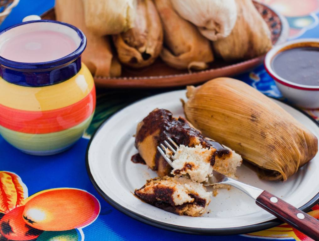 Tamales mexico, tamale with mole sauce, mexican food, traditional meal in Mexico. The tamale is sitting on a white plate with a fork. The plate is on a blue tablecloth surrounded by traditional Mexican pottery. Concept for the Best Tamales in Las Vegas.