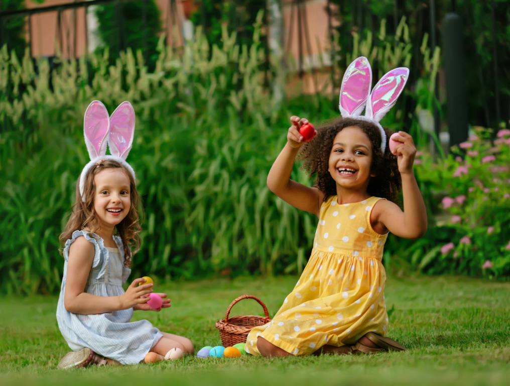 Two girls in Easter dresses and ears sit on a lawn holding plastic eggs and other Easter basket stuffers. They are wearing Easter dresses and bunny ears and smiling at the camera.