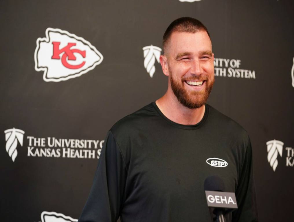 Travis Kelce wearing a black t-shirt and addressing a crowd after a game, with Kansas city Chiefs logo behind him.