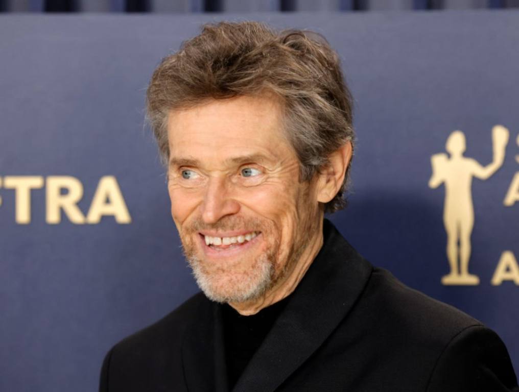 Willem Defoe poses for a photo in front of a blue Screen Actors Guild backdrop. He is smiling in a black collared shirt.