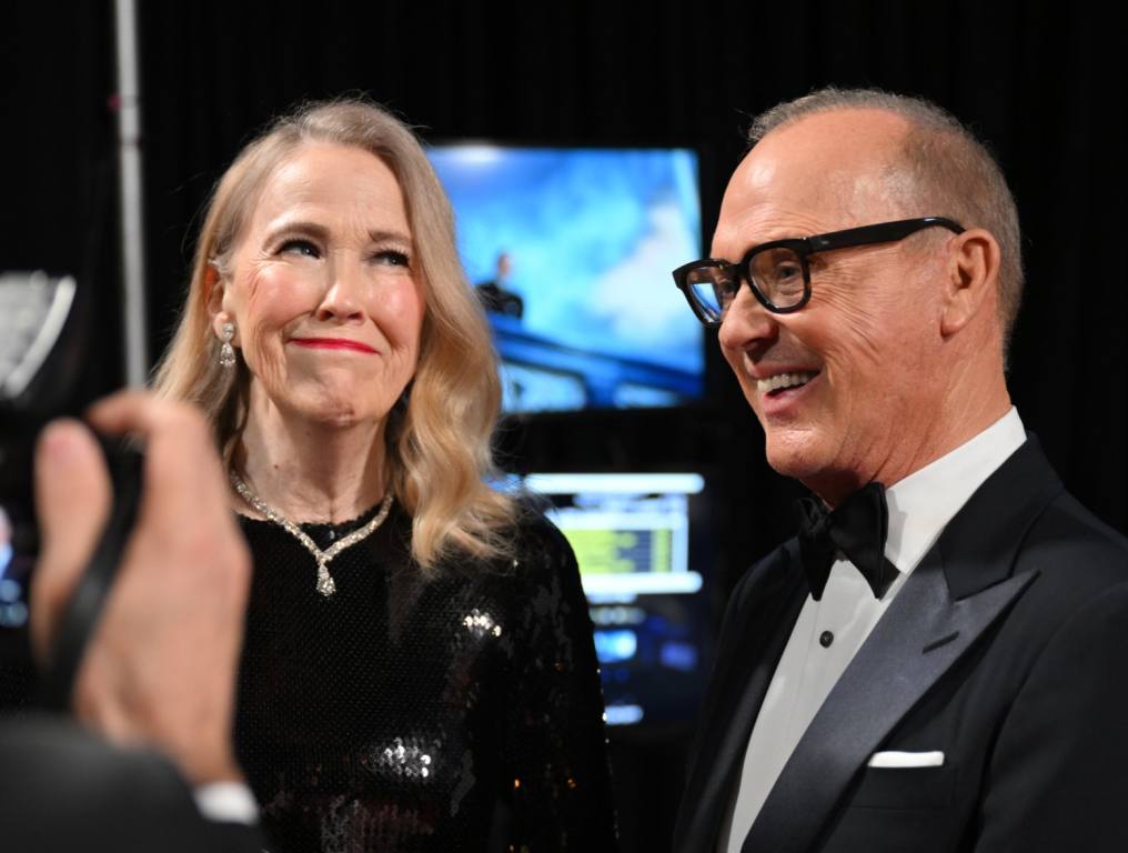 Catherine O'Hara and Michael Keaton, stars of Beetlejuice 2, are chatting backstage. O'Hara is in a black dress and Keaton is wearing a black and white tux and black glasses.