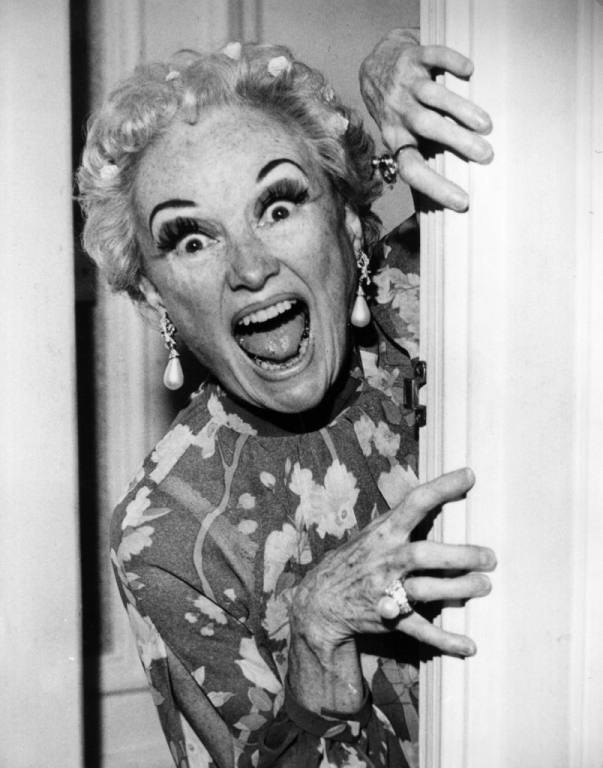 Black and white picture of Phyllis Diller looking silly from the side of a door.