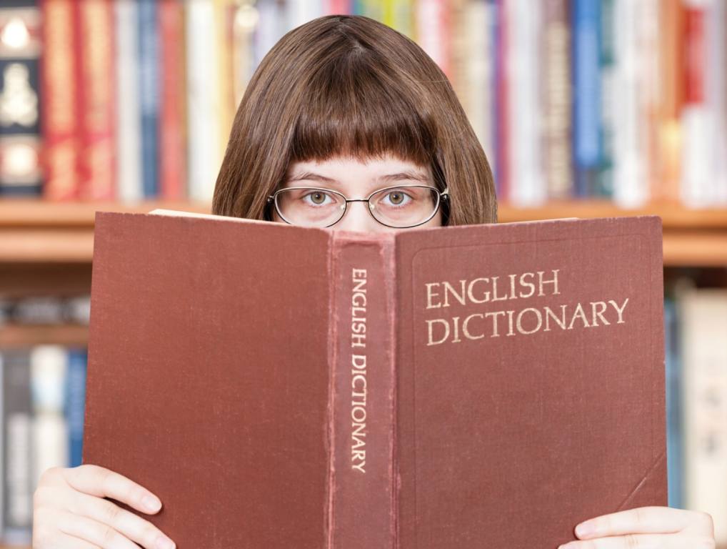 A young brunette with glasses peeks over the top of an English Dictionary. Many of the foreign words in the English language are mispronounced by Americans.