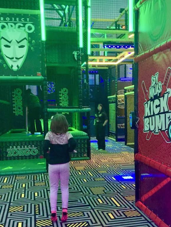 A young girl in white leggings and a black and white shirt is facing away from the camera as she looks at a room full of climbing jungle gyms. The floor is colorful. On one of the jungle gyms is a white mask with sinister looking face with a handlebar mustache.