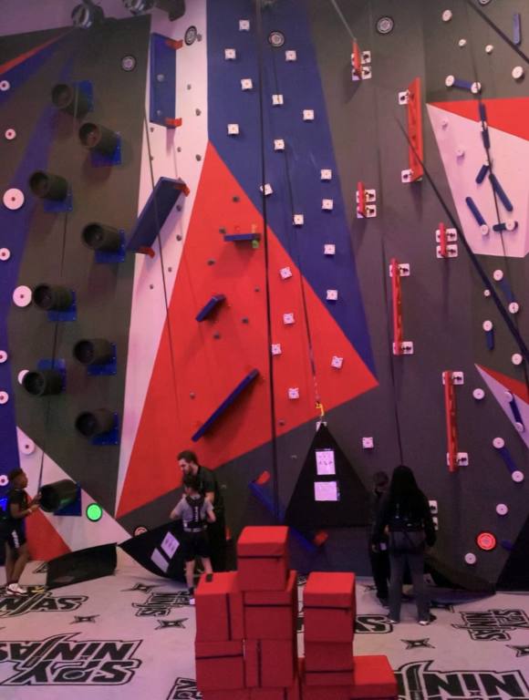 A room full of climbing walls. Red, blue and white geometric shapes on a gray wall. There are various obstacles and black climbing holds.