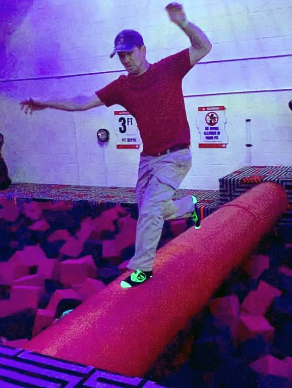 A man in khaki pants and a red shirt runs across a red vinyl spinning wheel in a foam pit at an indoor theme park. He has his arms out to balance as he tries not to fall off the wheel. The foam pit is filled with red and charcoal gray foam cubes.