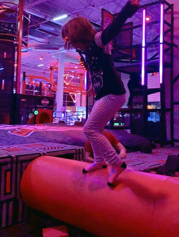 A young girl in white leggings runs across a giant spinning wheel in a theme park foam pit. The wheel is red vinyl and you can see the girl is in motion as she approaches the safety platform.