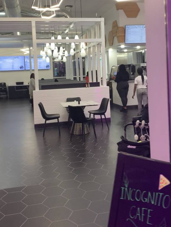 The entrance to a cafe shows a sign that reads "Incognito Cafe'. Beyond which you can see a gray floor, white cafe tables and dark gray chairs. There are people standing at the counter ordering food.