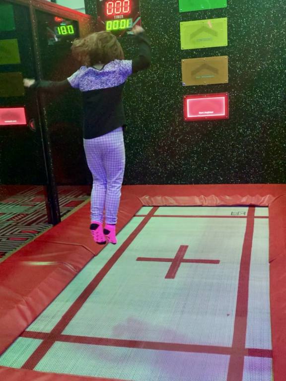 A female child in white leggings and black and white shirt is jumping on a large trampoline. She is facing away from the camera.