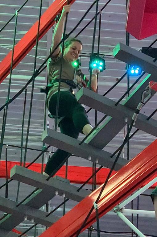 A woman in black leggings and a brown shirt walks across a gray suspended bridge. She is in a harness and holding on to the ropes that make up the sides of the bridge.