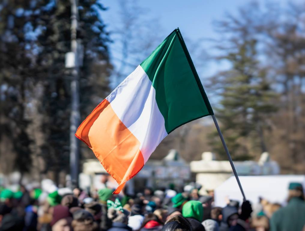 Interesting facts about St. Paddy's Day myths and folklore are revealed in this article. Photo of a St. Patrick's Day celebration. The center focus is on an Irish flag being held up above the crowd on a stick or pole on a sunny day at a large festival or parade. The bottom of the photo shows the heads of hundreds of people at this gathering. There are tall trees in the distance. The Irish flag is orange on the outer side of the rectangle, white in the middle, and green nearest the stick or pole.