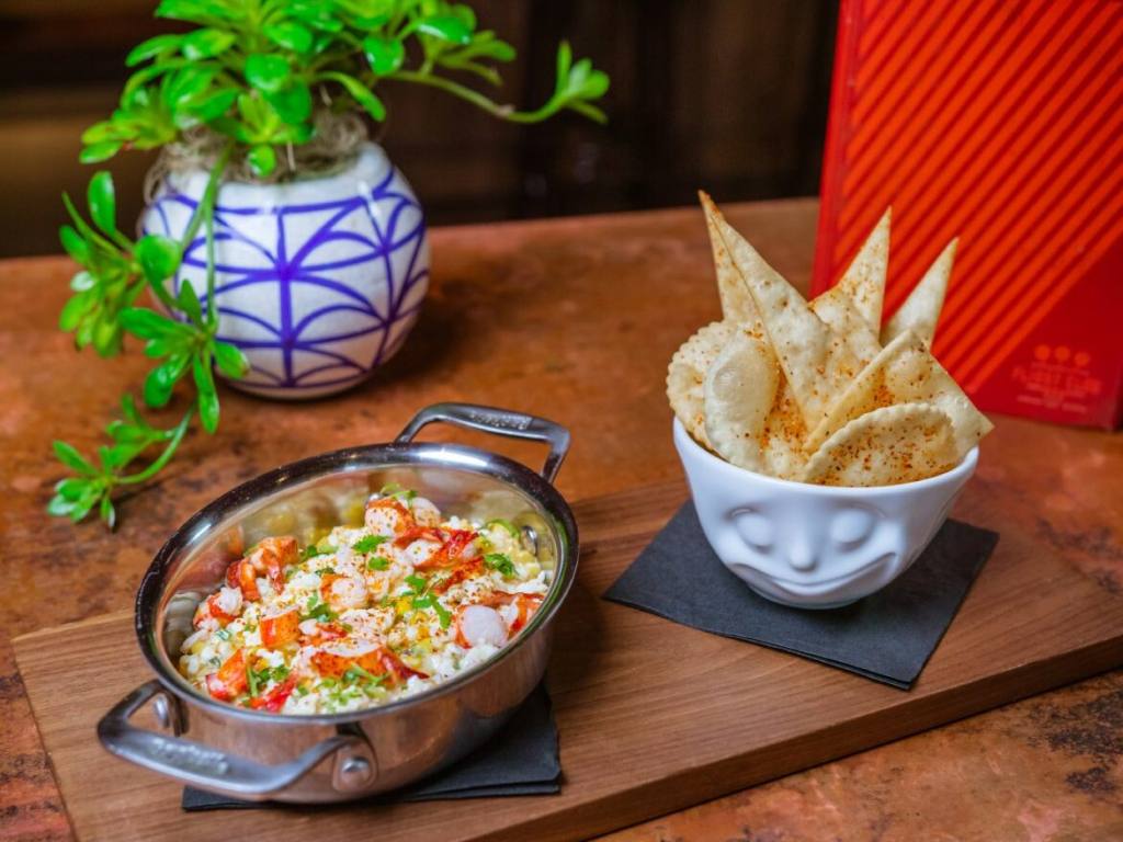 A dish of lobster elote dip sits on a wooden cutting board next to a bowl of chips and a potted plant. The Elote dip is one of the menu options at Flight Club Las Vegas.