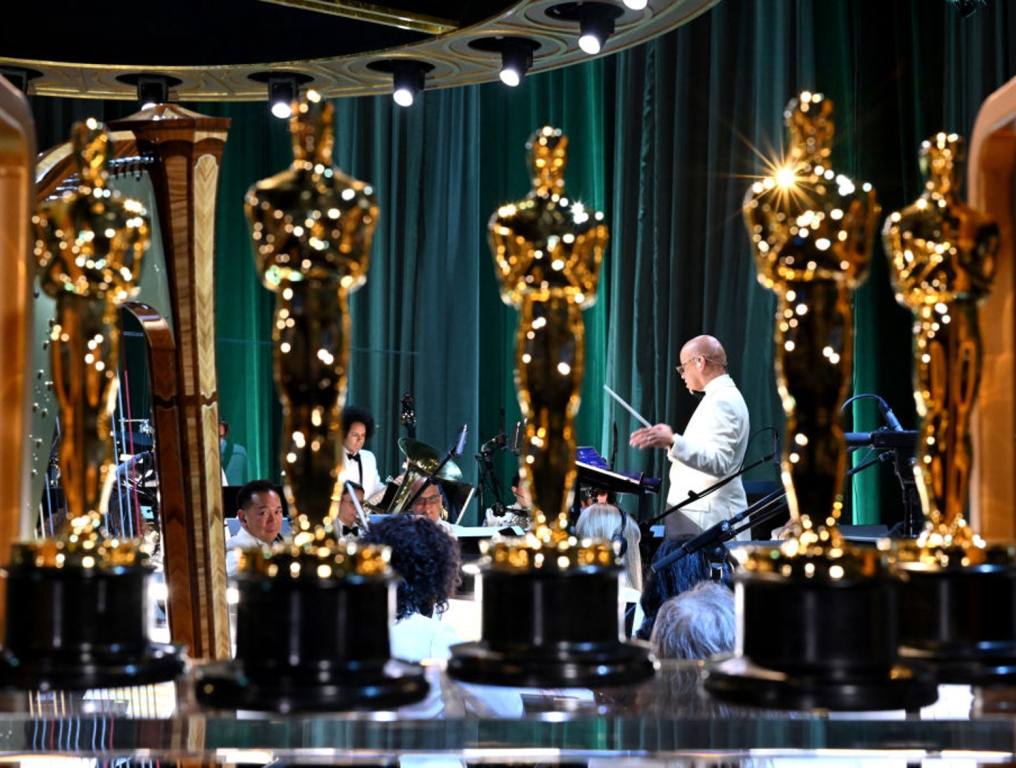 Oscar winning songs and popular music. Photo of five shiny, gold, Oscar statuettes displayed on an acrylic tabletop, backstage, at a recent Academy Awards ceremony. Behind the statuettes, an orchestra conductor stands on a pedestal, hold a baton in his left hand, and readies a seated orchestra to begin playing music. There is a forest green velvet curtain backdrop behind the orchestra's musicians.