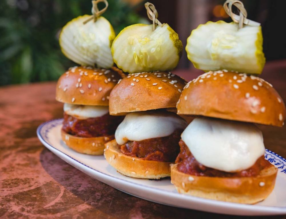 Three meatball sliders from Flight Club Las Vegas are presented on a white plate with a pickle skewered on the top of each. The plate is sitting on a wooden table.