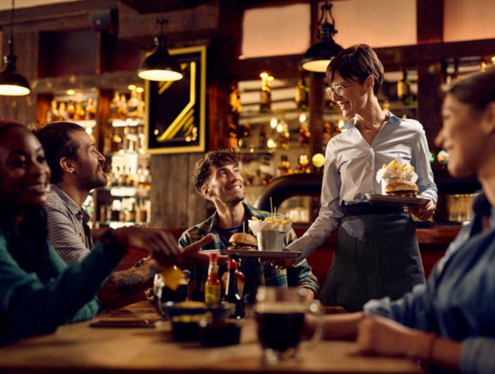 Happy waitress serving hamburger with French fries to group of guests in a bar, dining out concept