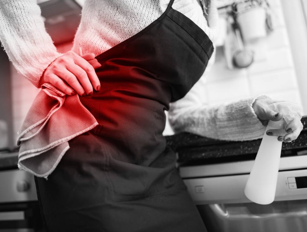 Health tips to help you avoid injuries when you're doing your spring cleaning. Photo of the torso and hips of a woman wearing a white sweater and a black apron. She is holding a plastic spray bottle in her left hand and a microfiber cloth in her right hand. Her right hand is also placed right above her right hip on her back as if it is in pain. This is a black and white photo with only the area of her back pain highlighted in red.