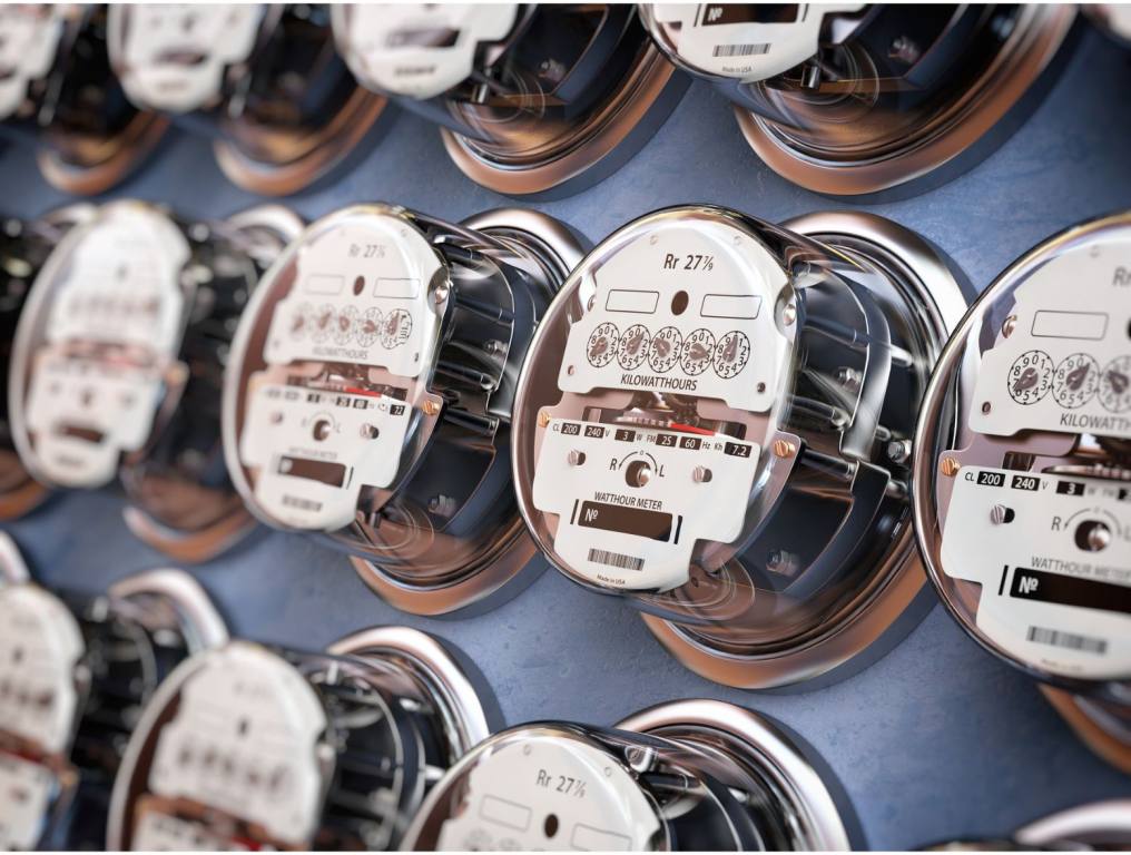 Close-up photo of a bank of electric meters representing high electric bills for Nevadans. Photo taken during daylight hours. Three full electric meters are visible in the middle of the photo. There is also a partial view of a row of electric meters above, and below, the middle row of electric meters. Each meter has a silver metal face on which a meter reader can determine how much energy is being used. There is a glass plate surrounding the silver metal facing so no one can tamper with the meter, but the meter reader can see through the glass.