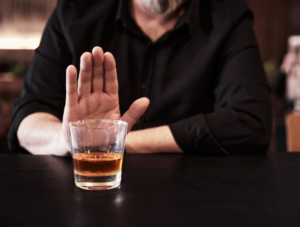 Man going sober right before St. Paddy's Day. Photo shows a man dressed in a black, V-neck shirt or sweater, holding up his right hand as if to say "No" to a shot of brown liquor in a glass set in front of him on a black tabletop at a pub, tavern, or bar.