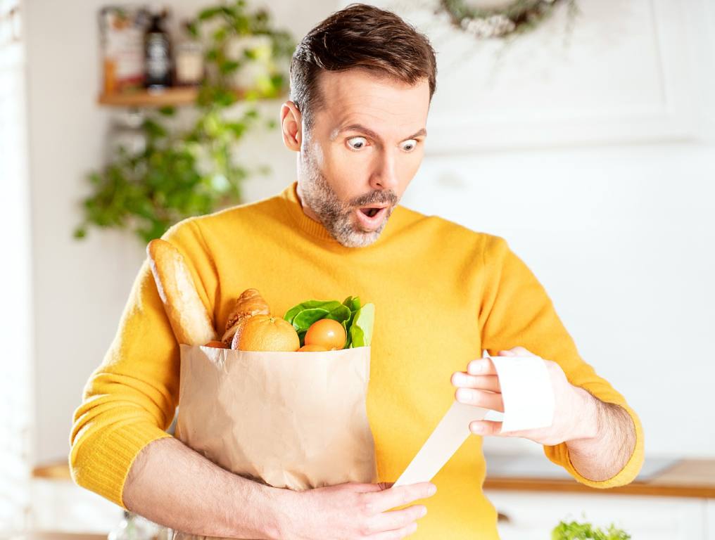 Nevadans are dealing with very high grocery prices. Photo shows a man in his 30's with a shocked and surprised look on his face. He is staring at his grocery store receipt, while holding a paper bag filled with groceries. A baguette, a grapefruit, an orange, and green leaf lettuce can be seen above the top rim of the grocery bag. The man is wearing a bright, mustard yellow sweater with the sleeves pulled up to the middle of his forearms. He has fairly short, combed and styled, reddish-brown hair and he is sporting a close-cropped gray and brown moustache and beard. He appears to be standing in his home, with bright sunshine streaming through the windows behind him. There is also a hanging plant basket behind his right shoulder.