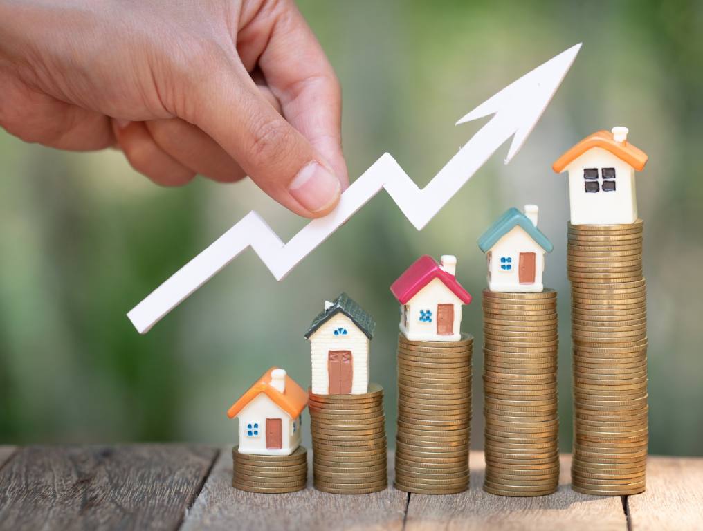 Photo depicting Nevada home values increasing over the past 10 years. Photo shows toy houses placed on top of five different stacks of coins. The stacks become larger from left to right. A human hand holds a white arrow which zig zags higher from left to right. It is above the stacks of coins with the toy houses on top. The stacks of coins with houses on top are placed upon a table made of wooden planks.