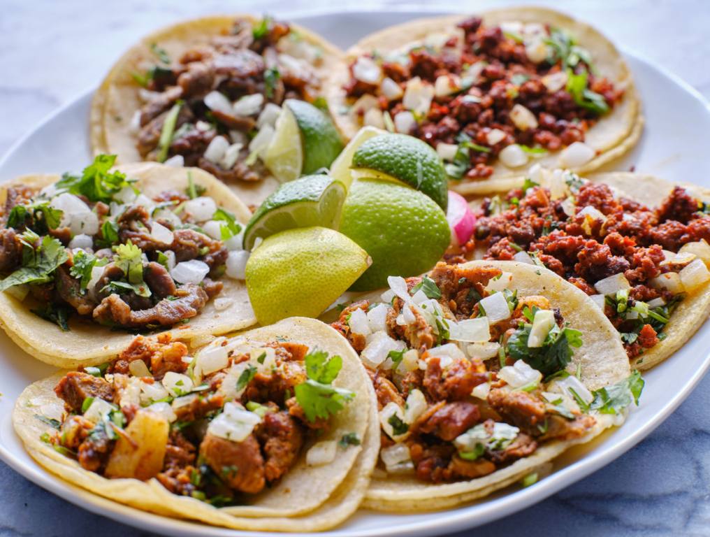 This image shows a white plate holding of three pairs of tacos. The pairs consist of carne asada, al pastor, and chicken tacos. Each taco consist of two corn tortillas, meat, cilantro, and diced onion. In the center of the plate sits a sliced up lime. The image is meant to depict tacos in Las Vegas.