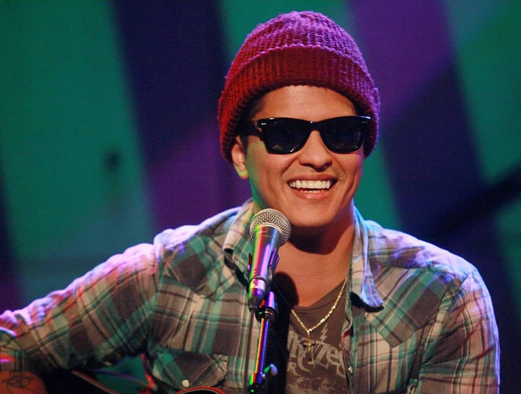 Bruno Mars wearing a maroon beanie and black sunglasses, while performing on a television show.