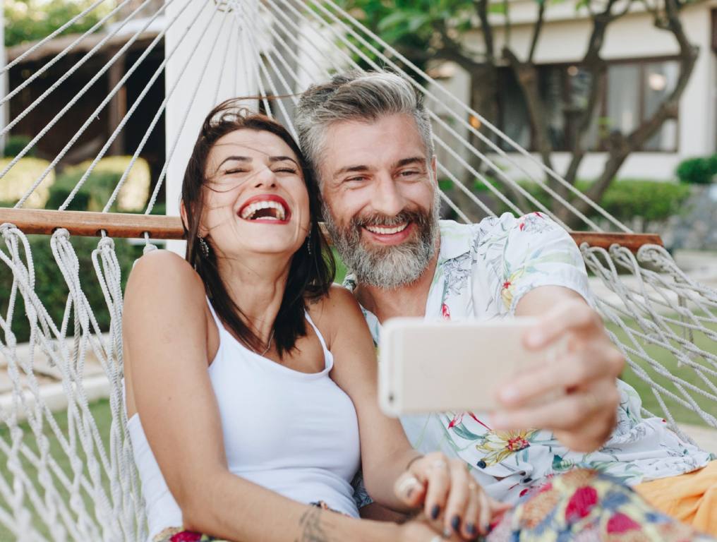 Living comfortably in Las Vegas is getting more expensive. Photo of a middle aged heterosexual couple is taking a selfie while laying in a white rope hammock. They are both laughing and smiling. The woman on the left has dark hair, red lipstick, and is wearing a white tank top. The man on the right has salt & pepper hair, beard, and moustache. He is hold a smart phone in his left hand to take the selfie photo. He is wearing an Hawaiian style "Aloha" shirt that is open at the collar. In the background, there looks to be a white building with brown trim.