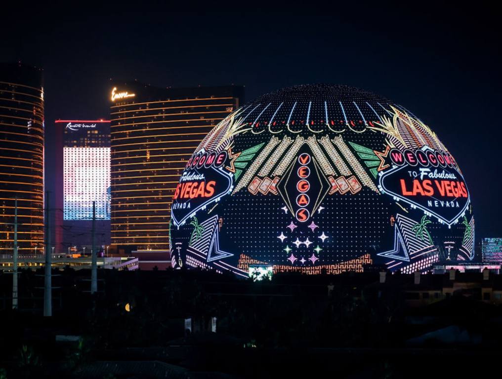 Nighttime photo of the Las Vegas Sphere where local art students may have their artwork displayed in the XO Student Design Challenge. The sphere shows various Las Vegas landmarks such as the "Welcome To Las Vegas sign." Other references to "Vegas" are also seen in bright lights on the sphere. To the left of the sphere, the outlines of Resorts World, Encore, and Wynn Hotels and Casinos can be seen.