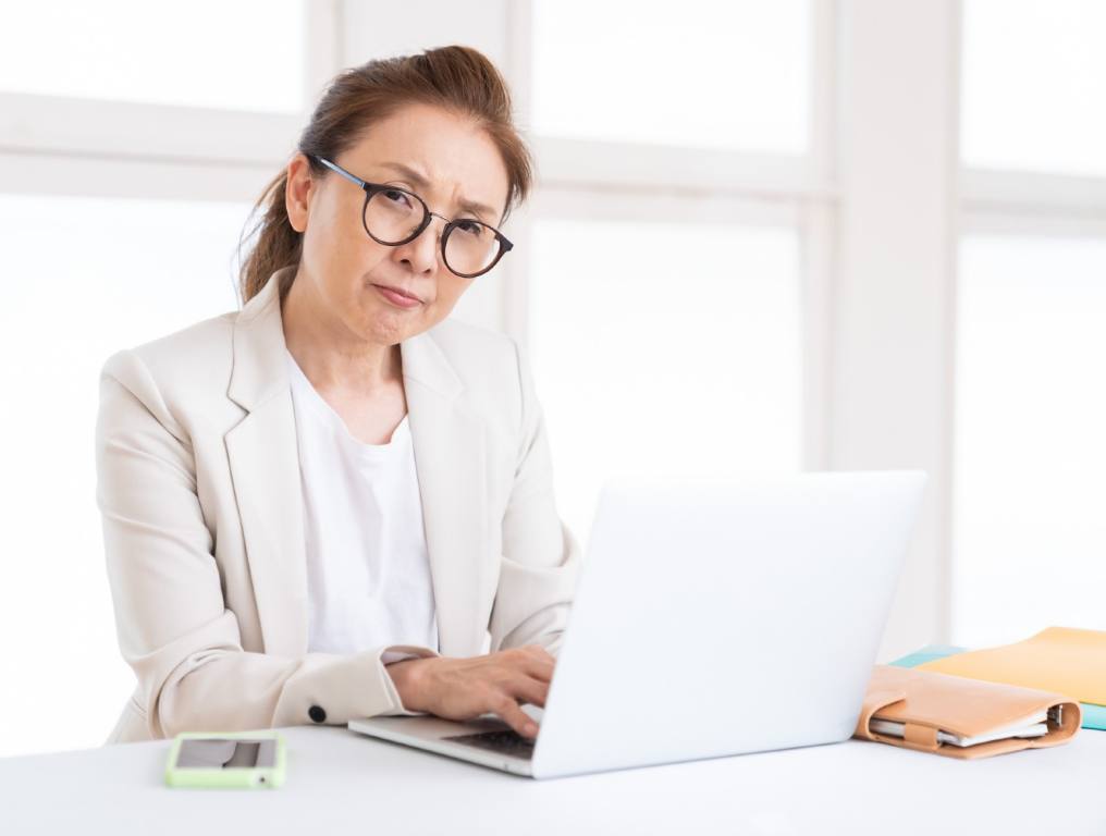 Photo depicting a woman unhappy with the gender pay gap in the technology sector. Photo shows an Asian, middle-aged, businesswoman who is typing on her white laptop computer. She is working on a white desk, in a mainly white room. There is light streaming through the four windows behind her. She has shoulder length brown hair pulled back away from her face. She is wearing large brown oval reading glasses. She is also wearing a white t-shirt underneath a white business suit jacket. On the table to her right is her mobile phone. On the table to her left is her date planner and some folders. She is looking directly into the camera and appears to be unhappy.