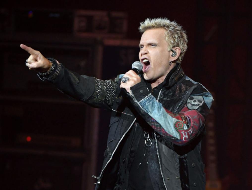 Music artist Billy Idol sings on stage while holding his finger up to the crowd. With his left hand, he holds his microphone to his mouth. He's wearing a black leather jacket.