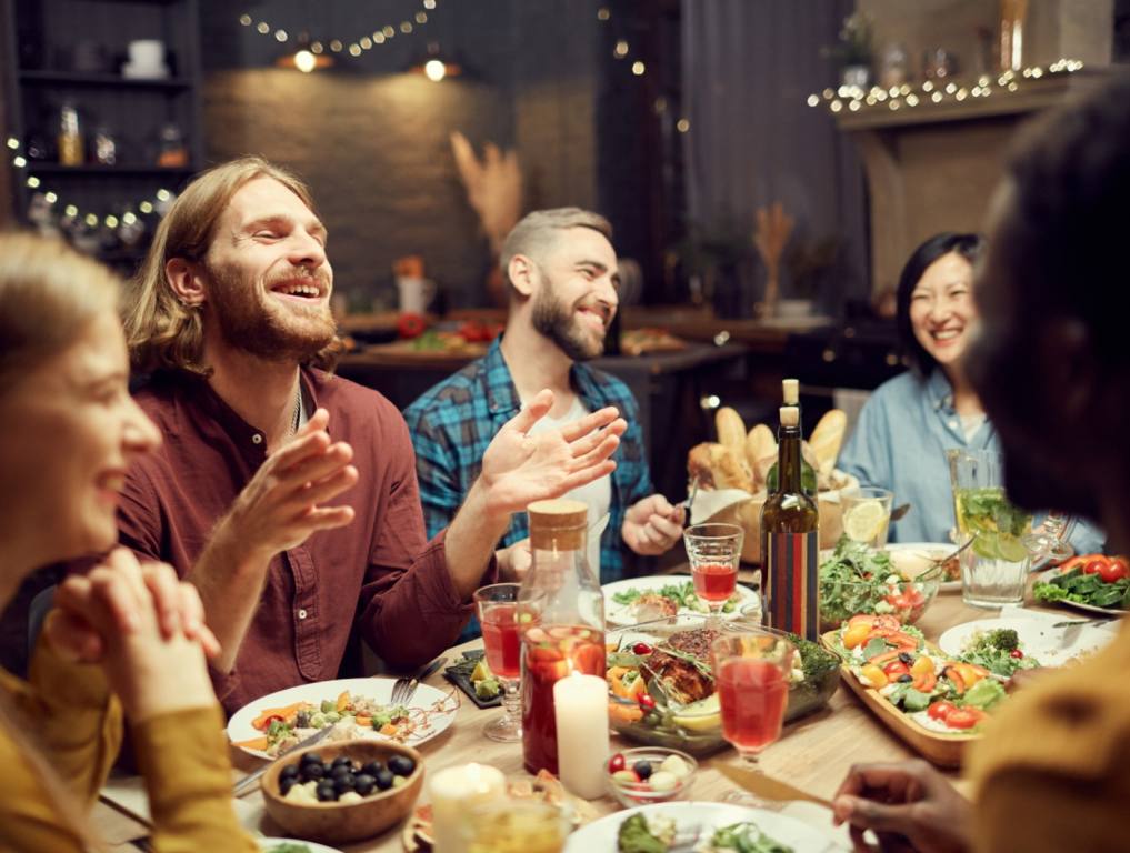 A group of young friends are sitting around a restaurant table in mid-conversation. They are laughing and eating various foods. New Henderson Restaurant concept.