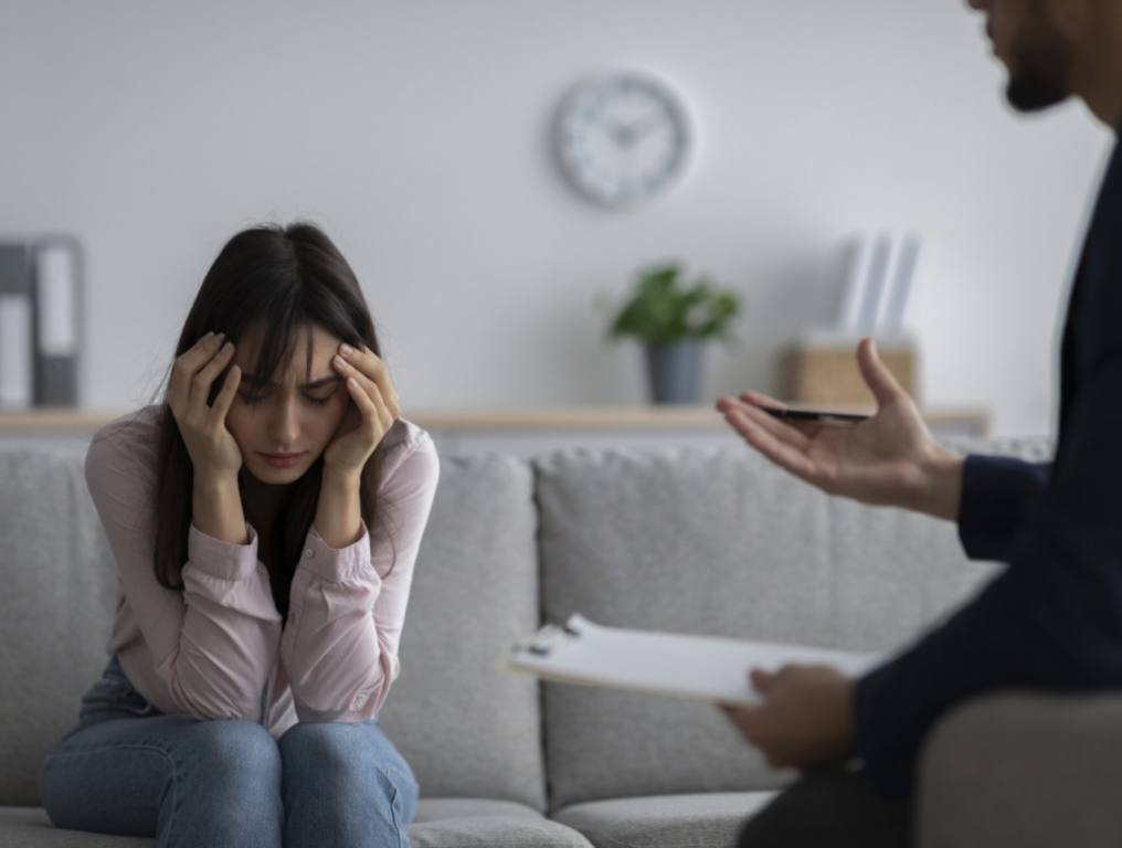 Depressed young woman touching head, seeking professional help with mental disorder at psychotherapist's office. Psychologist having session with desperate female patient. PTSD concept.