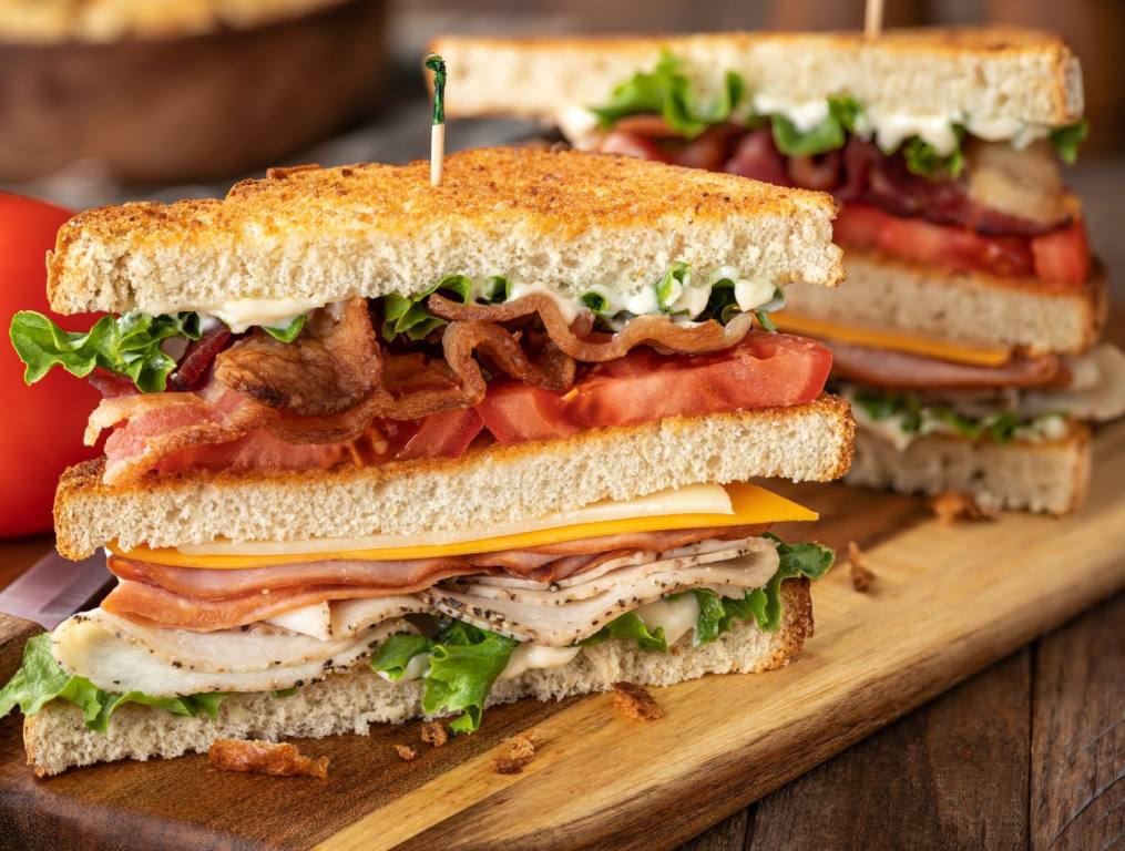 Club sandwich piled high with lettuce tomato bacon cheese and turkey. on toasted bread. The sandwich is cut in half on top of a cutting board