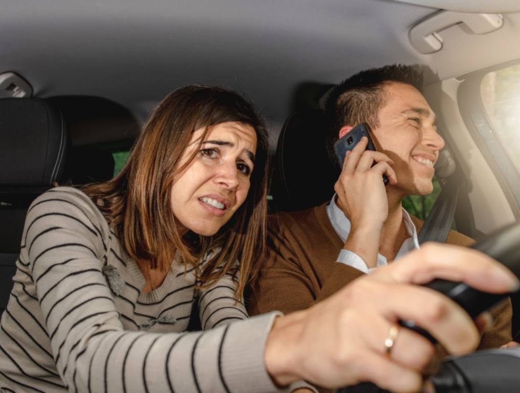 A man driving a car is talking on the phone and not paying attention. The woman in the passenger seat, noticing he is distracted driving, grabs the wheel with a worried look on her face.