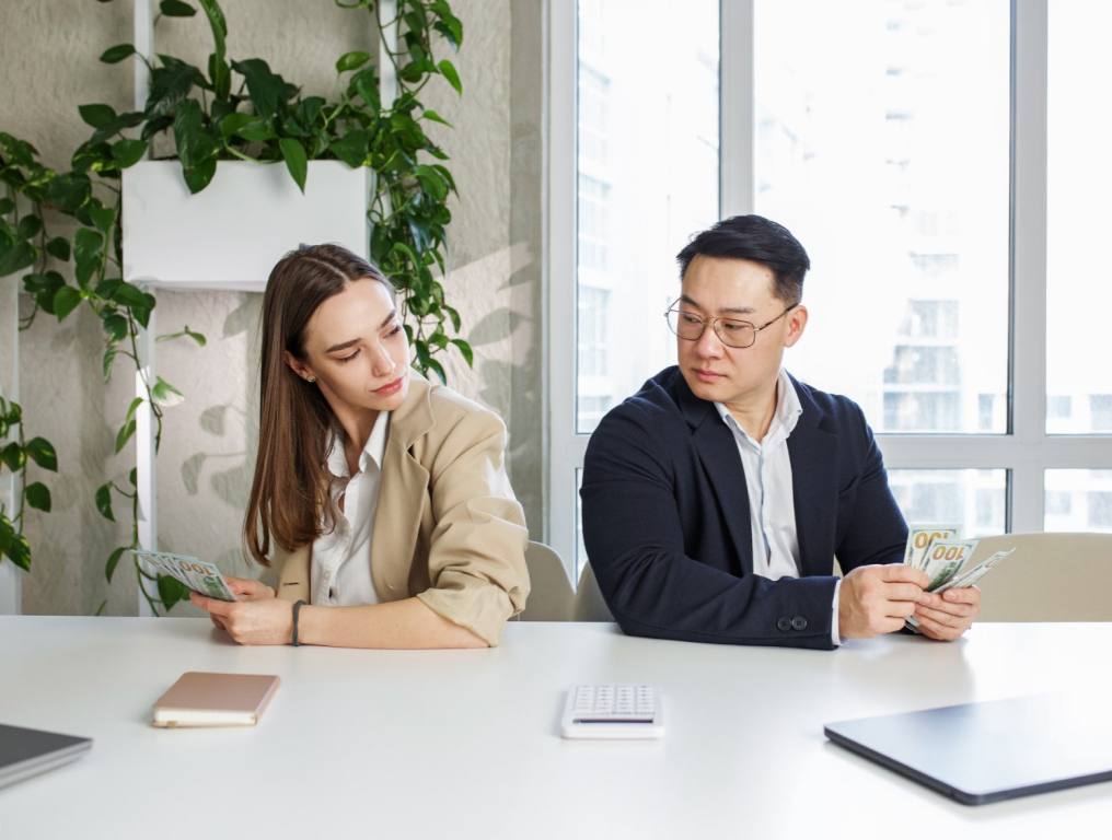 A white brunette woman sits at a white conference table next to an Asian business man. They are both holding cash and looking at each other suspiciously. Concept of gender wage gap.