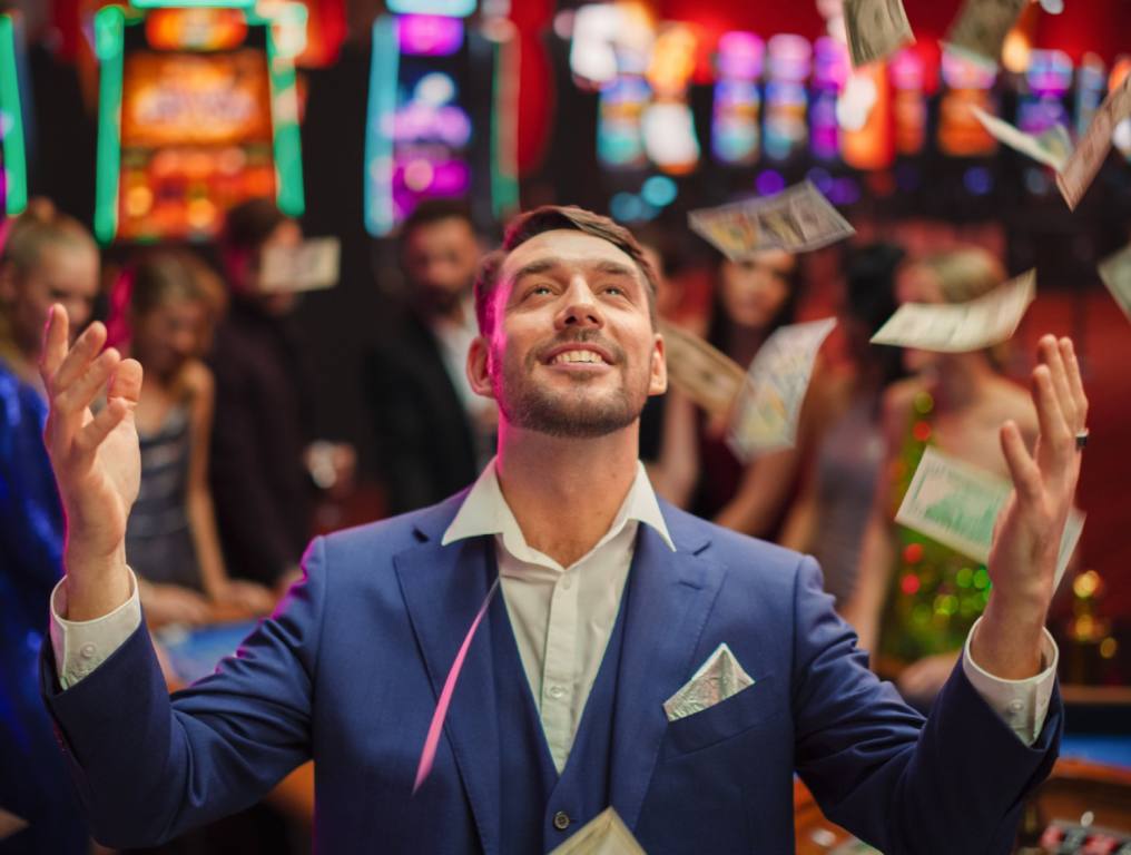 An excited young man in a blue suit is throwing his casino winnings up in the air. He is smiling and all the best casino games are in the background behind him.