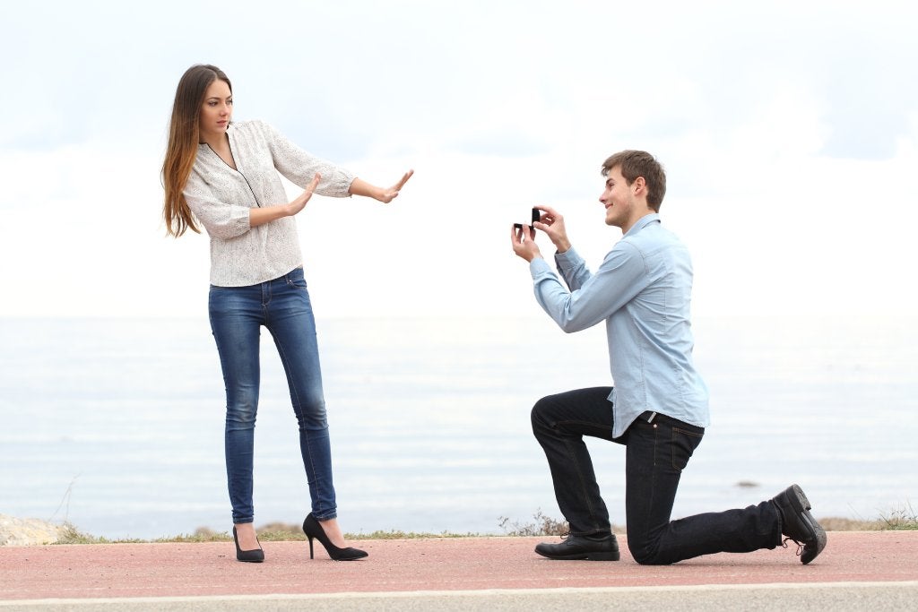 A light-haired brunette in a white shirt, jeans and heels is rejecting the proposal of a man suit pants and button up who is on his knees with a ring.