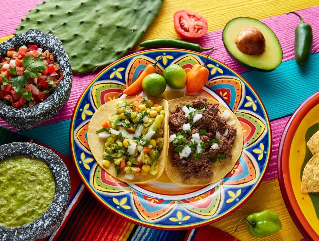 a table filled with Mexican foods. Salsa, guacamole, half of an avocado, limes and tomatoes. In the middle of the table is two tacos, the perfect food. Concept of the best tacos in Las Vegas.